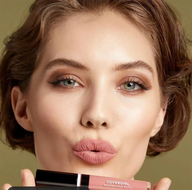 Beauty shot of a young woman with pouty lips holding CoverGirl Melting Pout Matte lipstick, soft focus, studio lighting on a neutral background.