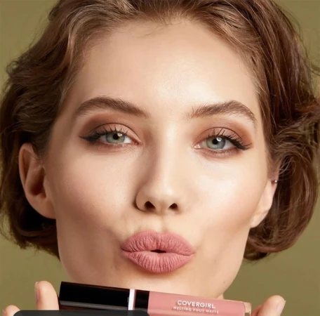 Beauty shot of a young woman with pouty lips holding CoverGirl Melting Pout Matte lipstick, soft focus, studio lighting on a neutral background.