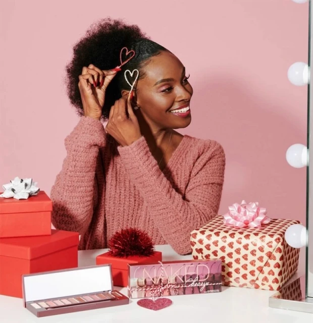 A smiling Black woman with heart hair clips poses indoors with gifts and makeup, bathed in soft pink light. Valentine's theme is present.