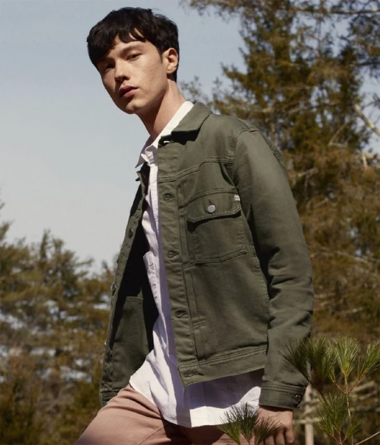 A young adult man in a green jacket and light pink pants stands outside in nature under a blue sky with some trees in the background.