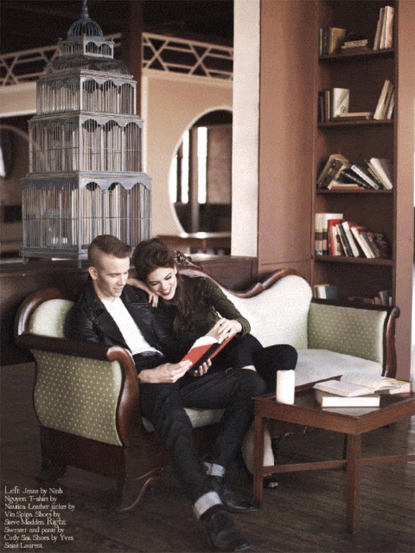 A couple sits on a light-green sofa in an indoor setting, reading together. The room has bookshelves and an ornamental birdcage.