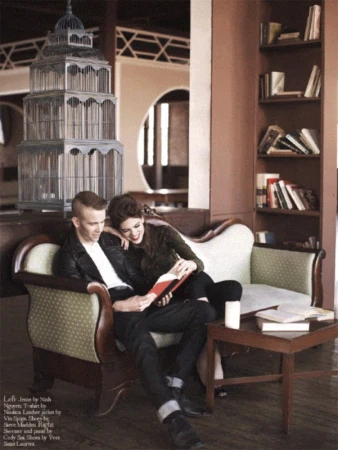 A couple sits on a light-green sofa in an indoor setting, reading together. The room has bookshelves and an ornamental birdcage.