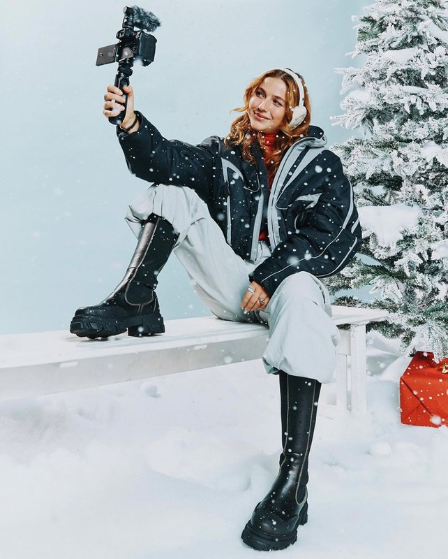 A woman in winter clothes sits on a bench in a snowy scene, recording a video with her phone. Christmas tree and gift in background.