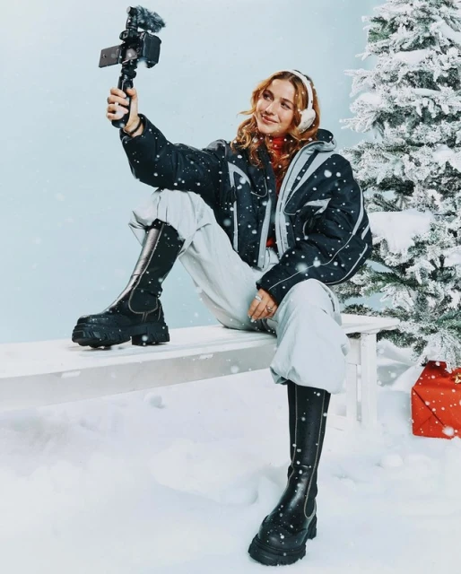 A woman in winter clothes sits on a bench in a snowy scene, recording a video with her phone. Christmas tree and gift in background.