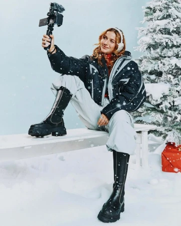 A woman in winter clothes sits on a bench in a snowy scene, recording a video with her phone. Christmas tree and gift in background.