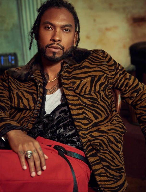 Portrait of a stylish adult male in an animal print jacket and red pants, posing indoors with a serious expression and warm lighting.