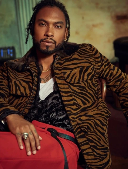 Portrait of a stylish adult male in an animal print jacket and red pants, posing indoors with a serious expression and warm lighting.