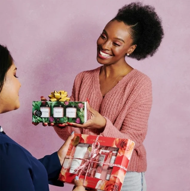 Two women exchanging beauty product gift sets. One woman hands a box to another, both smiling in an indoor setting.