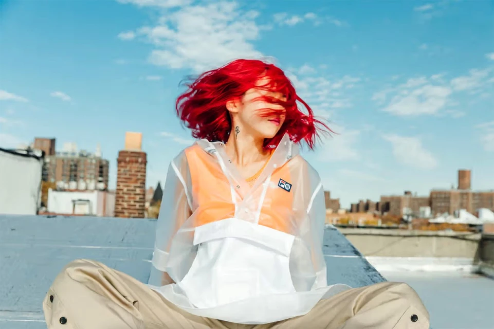 Young woman with vibrant red hair, wearing a transparent hooded jacket, sits cross-legged on a rooftop against a bright blue sky.