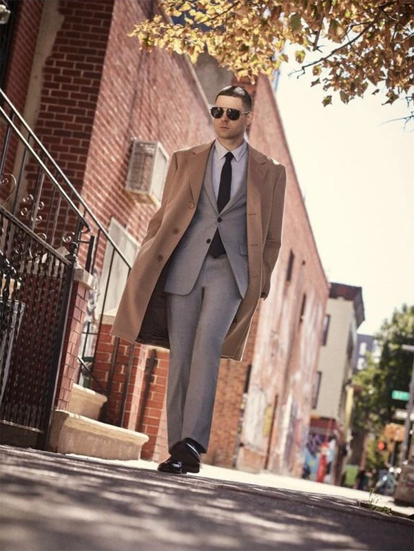Man in light grey suit and brown coat walking on a city street in sunglasses with brick buildings, trees, and a sidewalk.