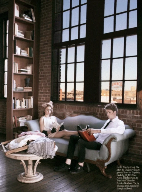 An adult male and adult female pose on a vintage-style settee in an industrial interior, reading, with a large window and bookshelf visible.