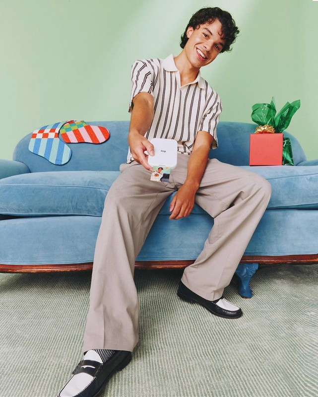 Young man sits on a light blue sofa with a small portable printer in his hand, gift box and colorful socks are in background.