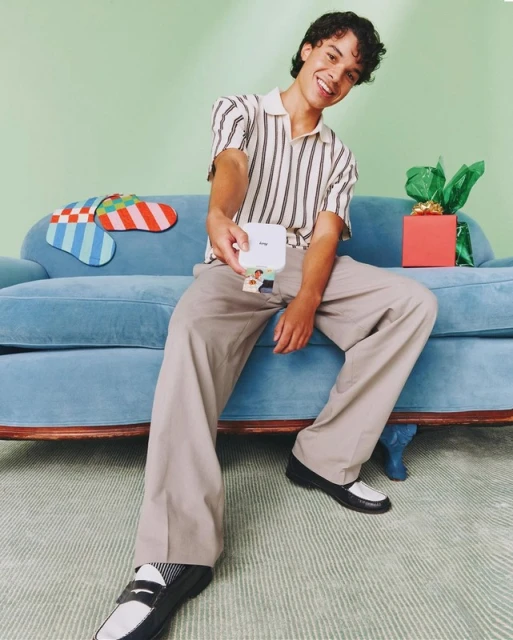 Young man sits on a light blue sofa with a small portable printer in his hand, gift box and colorful socks are in background.
