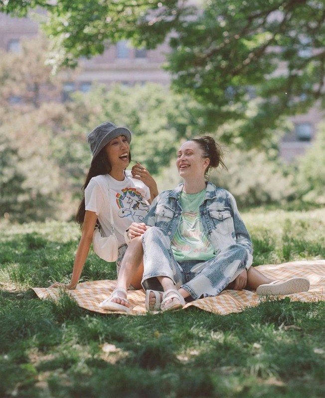 Two happy adult women sitting on a picnic blanket in a grassy park with trees and a building visible in the background on a sunny day.