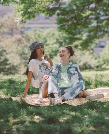 Two happy adult women sitting on a picnic blanket in a grassy park with trees and a building visible in the background on a sunny day.