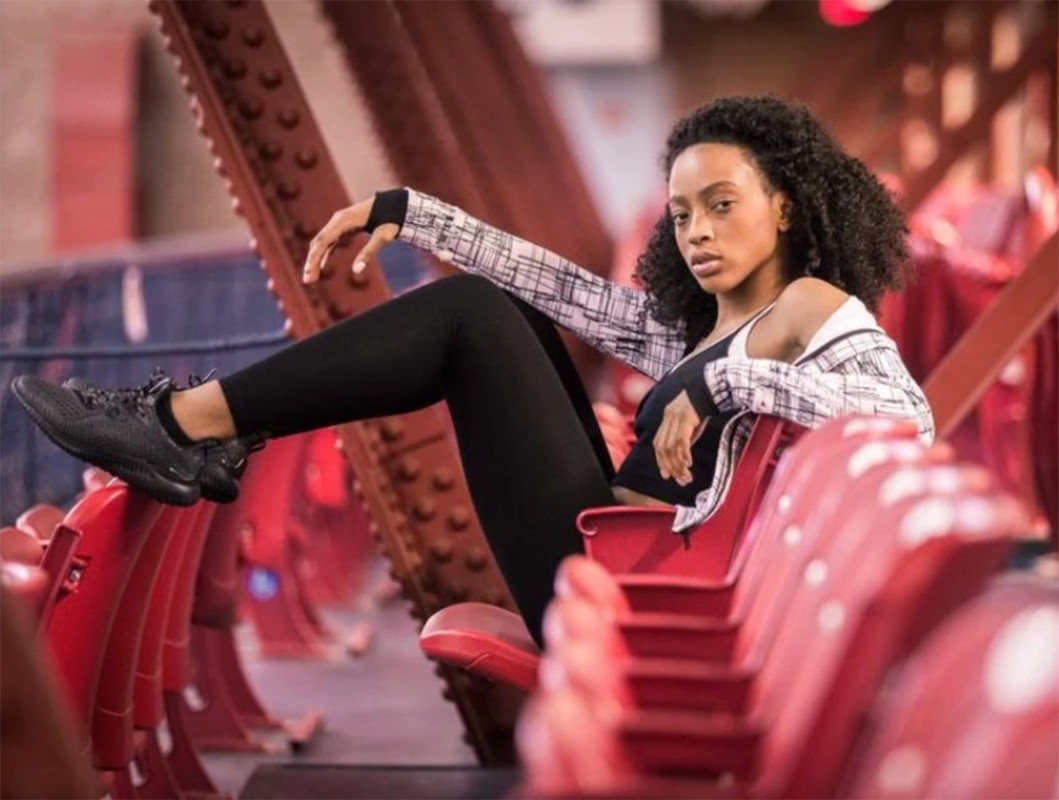 A young adult woman with curly hair poses in black athletic wear and a patterned jacket, sitting on red stadium seating in an urban setting.