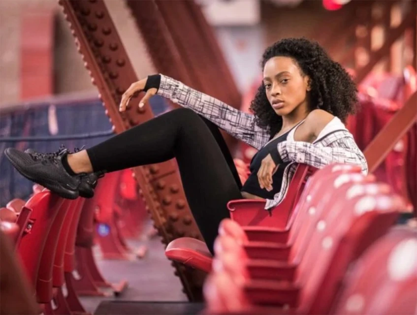 A young adult woman with curly hair poses in black athletic wear and a patterned jacket, sitting on red stadium seating in an urban setting.