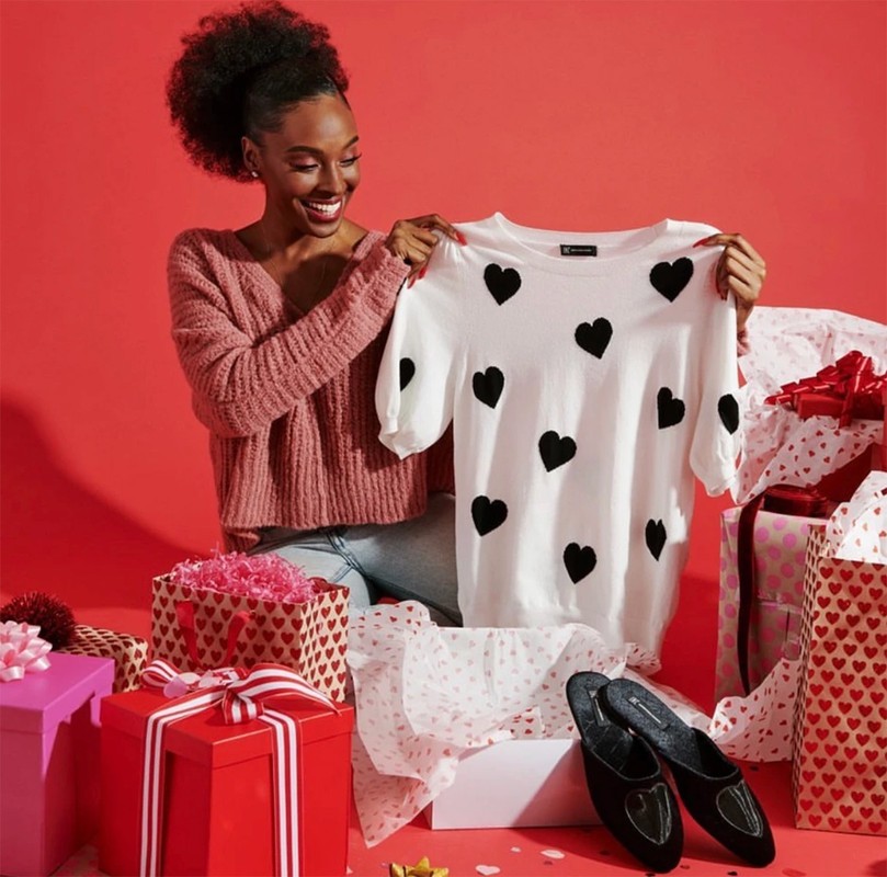 A smiling African-American woman is seated among gift boxes and holds up a white sweater decorated with black hearts in a bright studio.