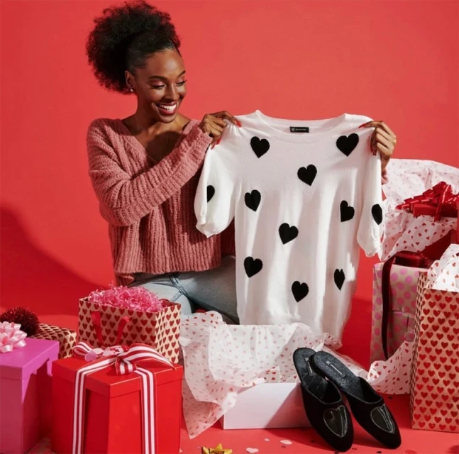 A smiling African-American woman is seated among gift boxes and holds up a white sweater decorated with black hearts in a bright studio.