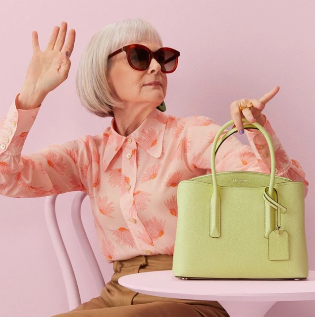 Elderly woman models a chartreuse satchel purse in a studio with a pink backdrop, wearing sunglasses and a patterned blouse.