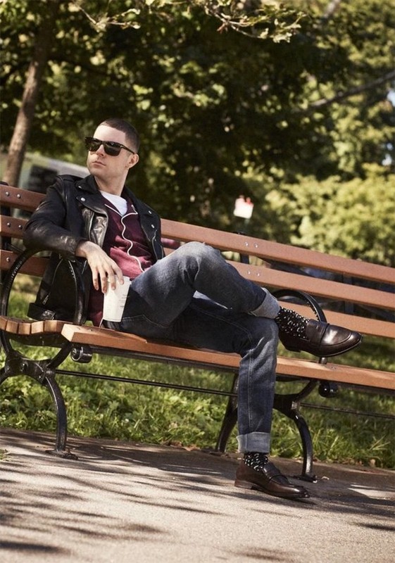 A stylish adult male is sitting on a park bench wearing sunglasses, a black leather jacket, and dark-wash jeans, relaxing outdoors.