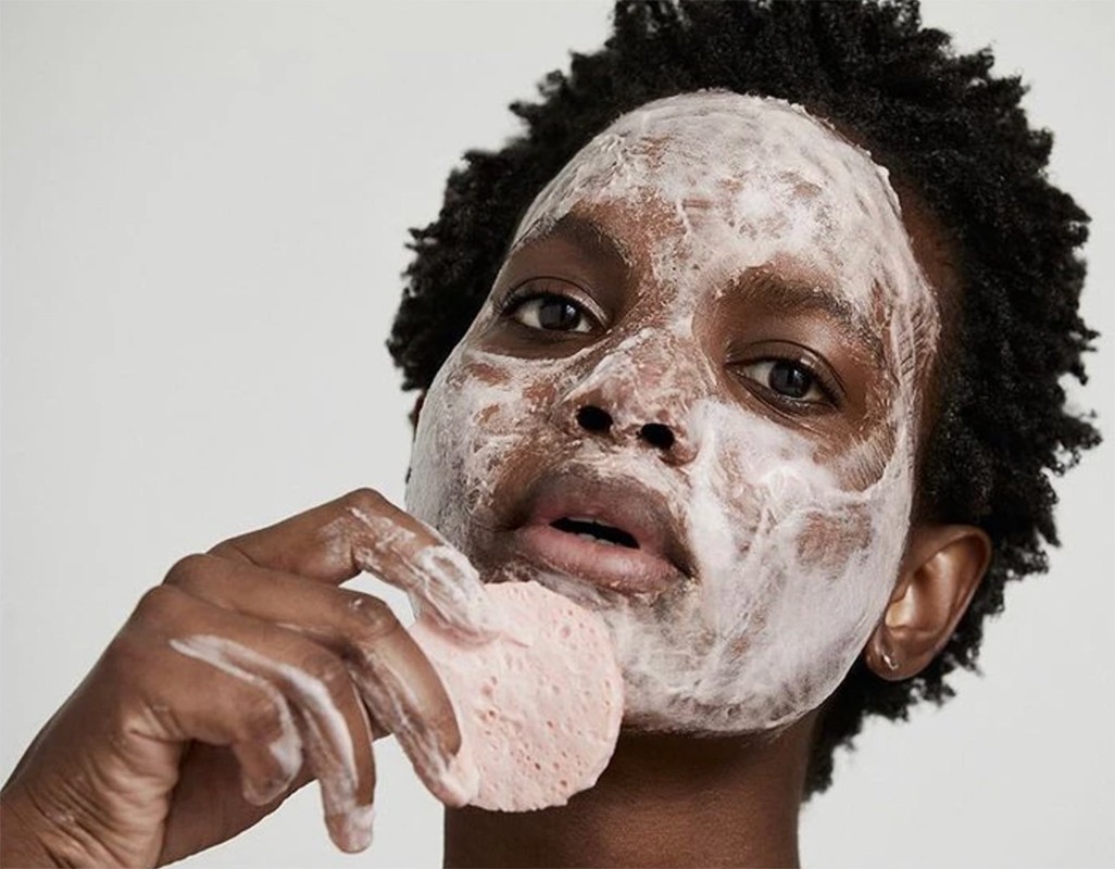 Close-up of a dark-skinned woman with white cream on her face, holding a pink sponge to her cheek in an indoor setting.