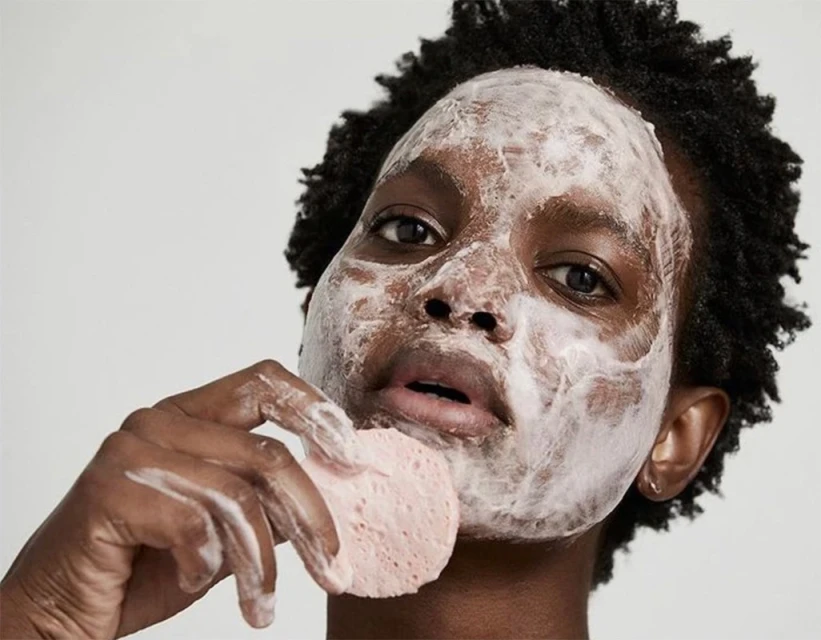 Close-up of a dark-skinned woman with white cream on her face, holding a pink sponge to her cheek in an indoor setting.