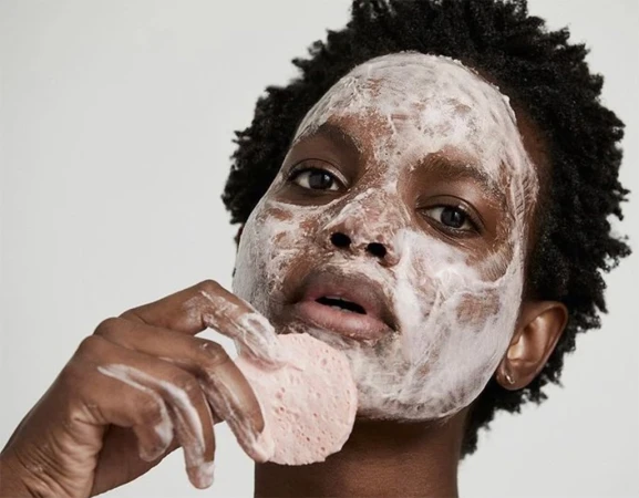 Close-up of a dark-skinned woman with white cream on her face, holding a pink sponge to her cheek in an indoor setting.