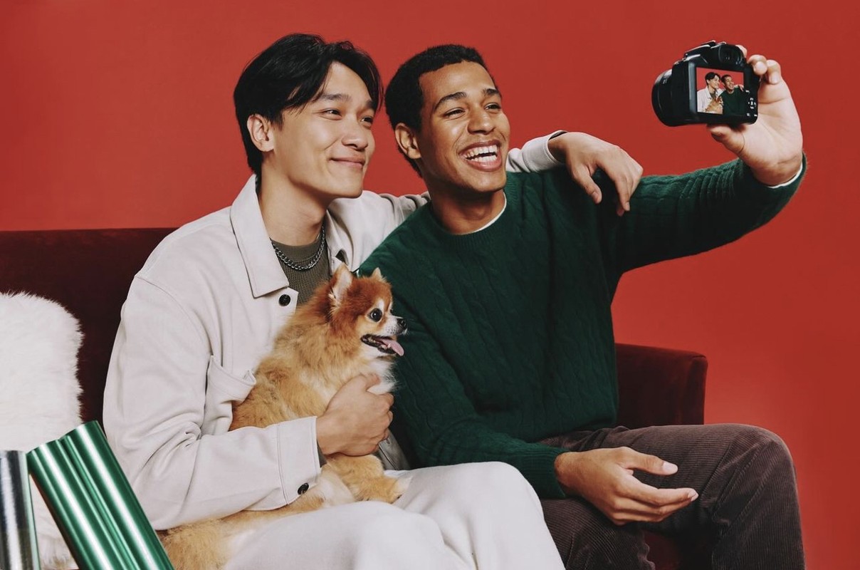 Two young men smile for a camera selfie while sitting on a red couch with a fluffy dog and wrapped gifts against a red backdrop.