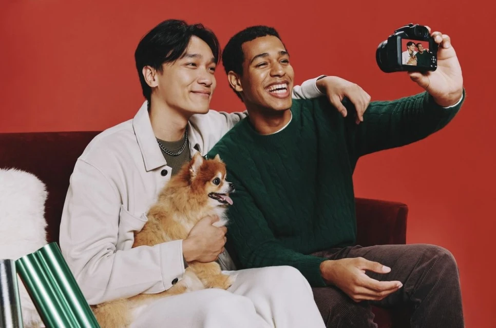 Two young men smile for a camera selfie while sitting on a red couch with a fluffy dog and wrapped gifts against a red backdrop.