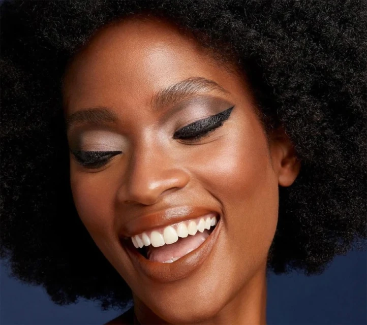 Close-up of a smiling African American woman with dark hair and eyeliner against a blue backdrop, showcasing beauty and makeup.
