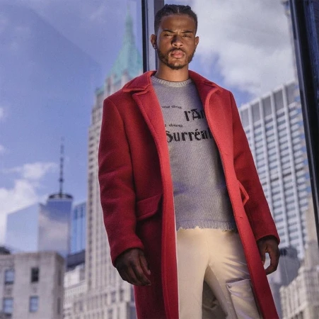 Black man with braided hair wearing a red overcoat and grey sweater against a skyscraper background
