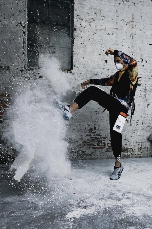 A person in a mask kicks a cloud of white powder in an urban environment with a distressed brick wall and window in the background.