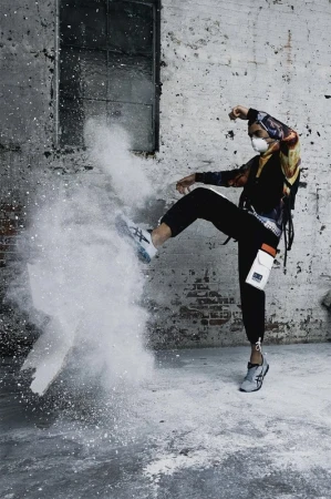 A person in a mask kicks a cloud of white powder in an urban environment with a distressed brick wall and window in the background.