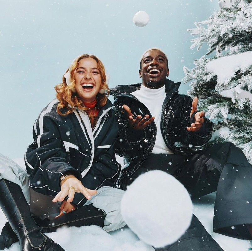 A laughing couple in winter coats sit in faux snow next to a Christmas tree, playfully tossing snowballs against a soft blue background.