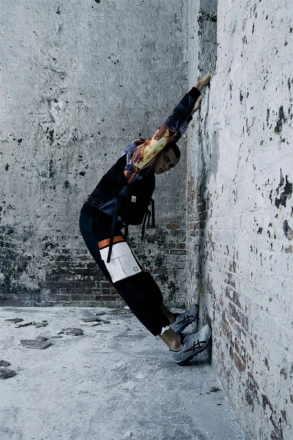 A person in athletic wear leans against a distressed concrete and brick wall, showcasing fashion in an urban environment with cool tones.