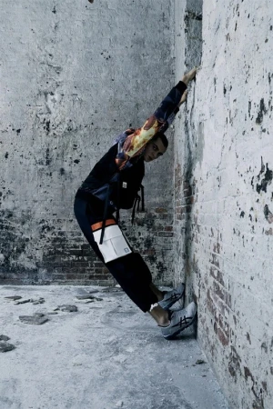 A person in athletic wear leans against a distressed concrete and brick wall, showcasing fashion in an urban environment with cool tones.