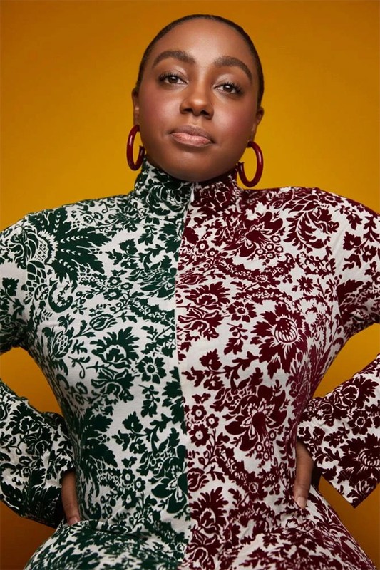 Portrait of a Black woman in a high-neck, patterned dress with a yellow background, red hoop earrings, and hands on her hips.