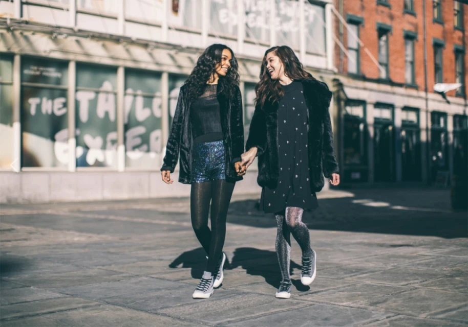 Two young women walking on a city street, holding hands, wearing fashionable outfits of dark colors and shiny tights, daytime.