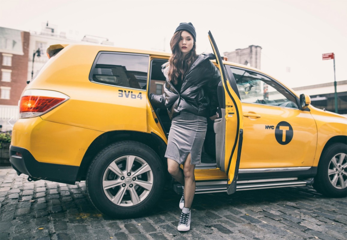 Young woman exiting yellow taxi in urban setting, wearing black puffer jacket and beanie, posing on cobblestone street.