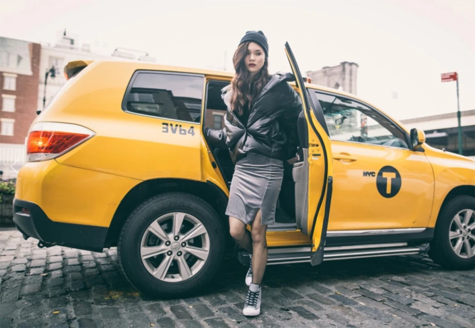Young woman exiting yellow taxi in urban setting, wearing black puffer jacket and beanie, posing on cobblestone street.