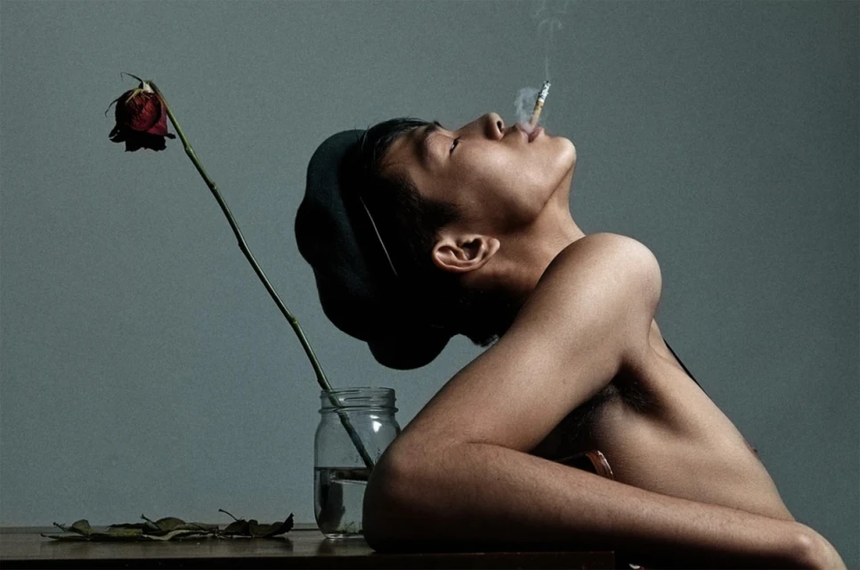 Adult with a cigarette poses next to a wilting red rose in a glass jar on a brown table in a moody, artistic portrait, lit with studio lighting.