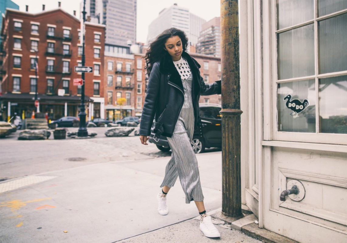 A woman walks on a city street wearing a black jacket and shimmering jumpsuit, with buildings and a street corner in the background.