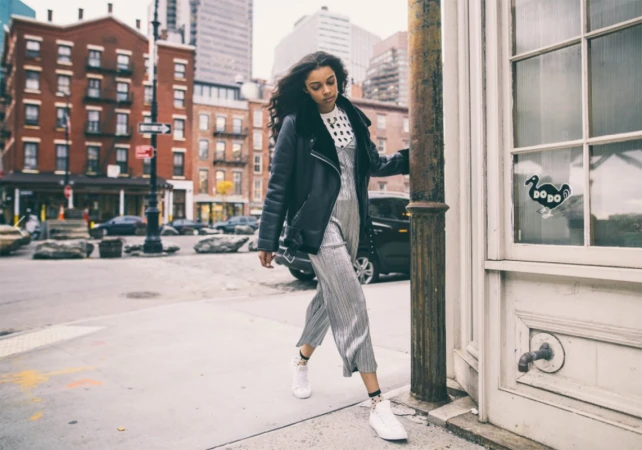 A woman walks on a city street wearing a black jacket and shimmering jumpsuit, with buildings and a street corner in the background.