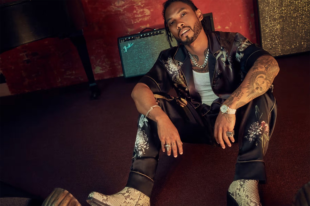 Stylish adult male with tattoos in patterned silk shirt and pants sits in photo studio next to amplifier, wearing jewelry and snakeskin boots.