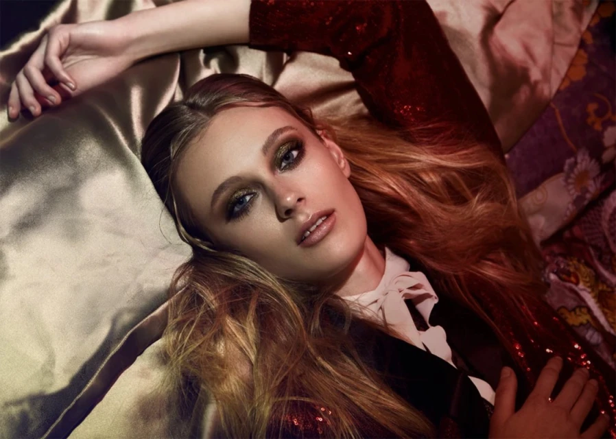 Close-up fashion portrait of a woman with long auburn hair lying on patterned fabric. She wears a sequin top and smokey eye makeup.