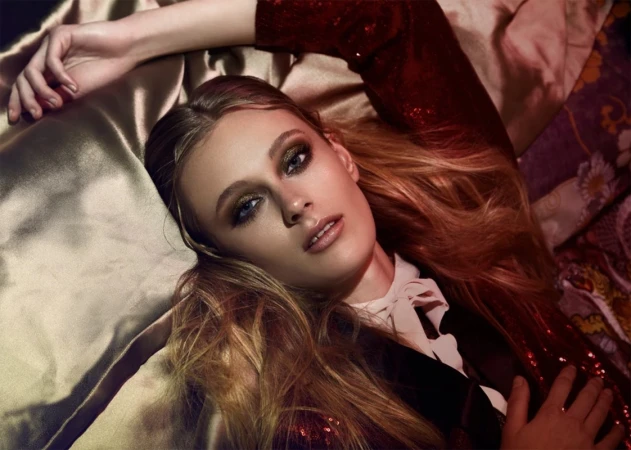 Close-up fashion portrait of a woman with long auburn hair lying on patterned fabric. She wears a sequin top and smokey eye makeup.