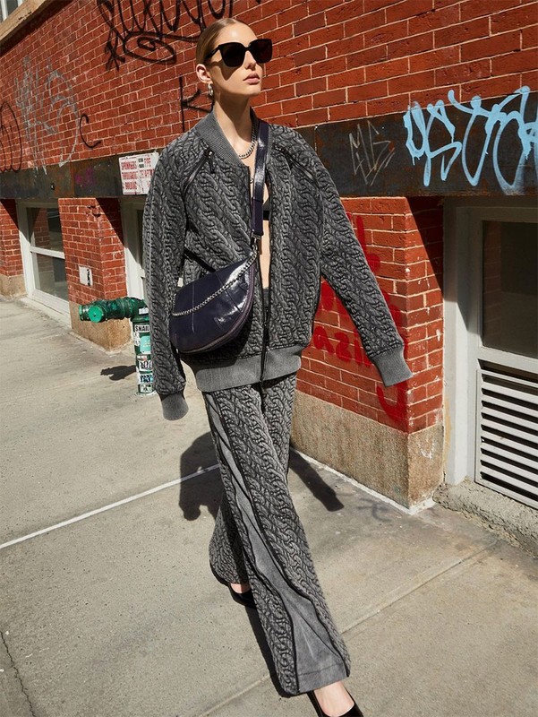 Adult woman wearing grey patterned jacket and pants with a crossbody bag walks along a city sidewalk with a brick building backdrop.