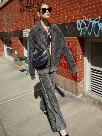 Adult woman wearing grey patterned jacket and pants with a crossbody bag walks along a city sidewalk with a brick building backdrop.