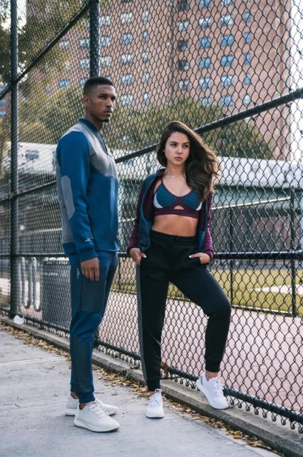 Athletic couple in workout clothes poses outdoors against a chain-link fence with an apartment building and trees in the background.